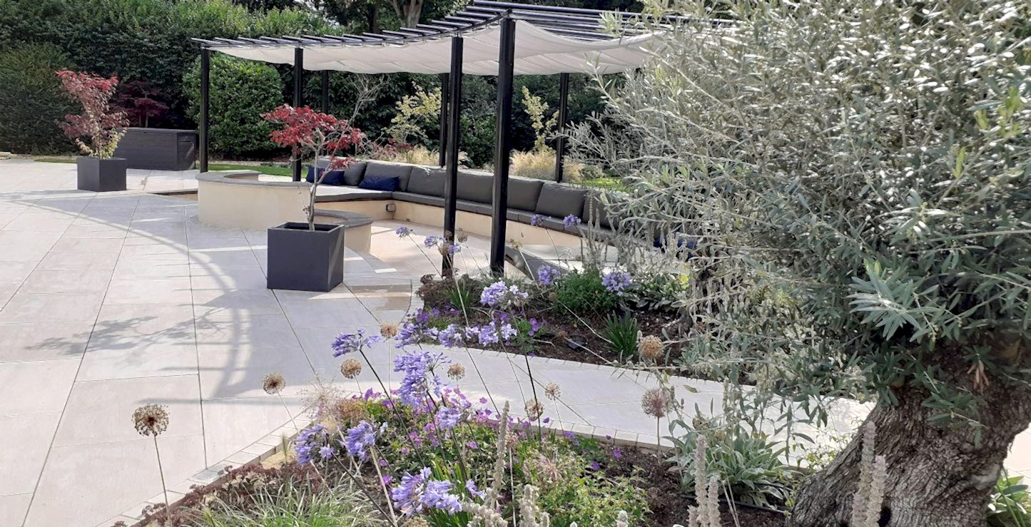 Garden design in Windsor, Berkshire with sunken seating area with built-in benches, outdoor fireplace, outdoor kitchen, outdoor shower, swimming pool and Mediterranean planting scheme.